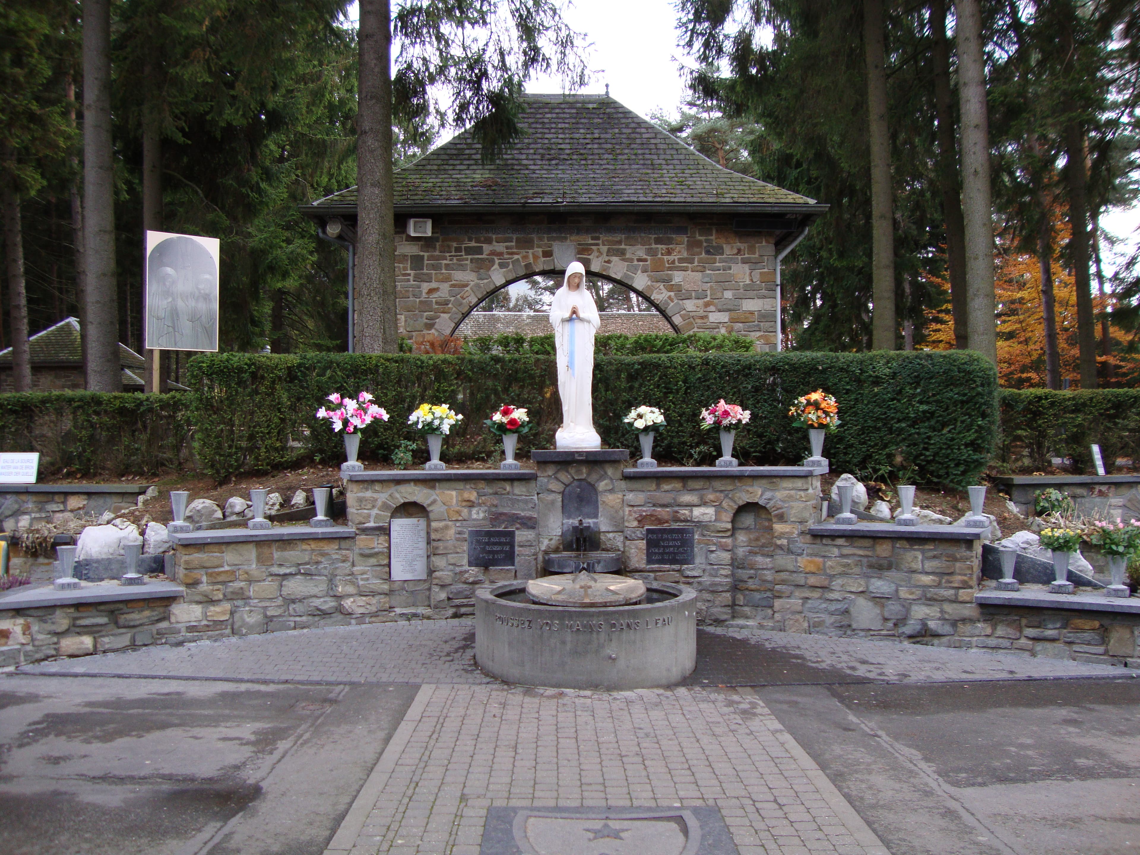 Source aménagée du sanctuaire de Banneux, entourée de fleurs et surmontée de la statue de la Vierge des Pauvres